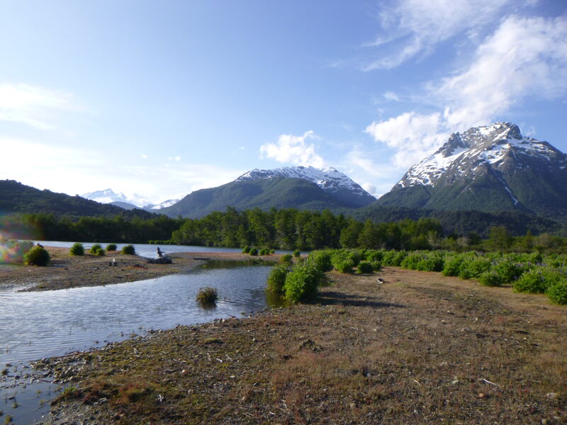 The image shows a scenic landscape with a river flowing through a valley. Lush green trees line the riverbanks, and in the background, majestic mountains rise towards the sky. The mountain peaks are covered with snow, contrasting with the greenery below. The sky is blue with scattered clouds, adding to the beauty of the natural scene.